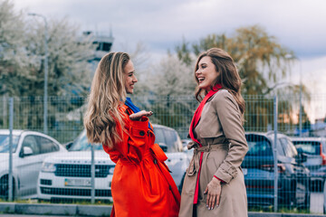 Fashionable happy smiling blonde woman wearing orange coat, blue jeanse and neckchain communicate with her girlfriend on the street.