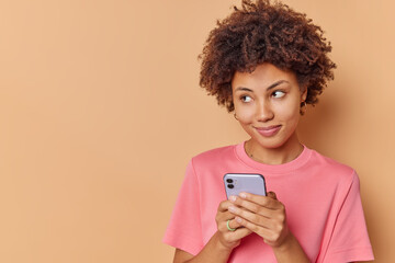 Pretty curly haired young woman with dreamy expression uses mobile phone sends text messages to friend dressed casually concentrated away poses against beige background blank copy space on left