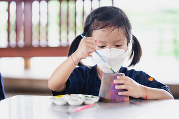 Kid making craft with paintbrush. Little girl wearing surgical white face mask to prevent spread virus and dust pollution PM2.5.