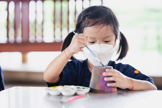 Kid making craft with paintbrush. Little girl wearing surgical white face mask to prevent spread virus and dust pollution PM2.5.