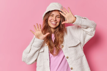 Horizontal shot of good looking young woman has playful expression raises palms smiles joyfully entertains you wearsfur jacket and hat poses against pink background. Positive emotions concept