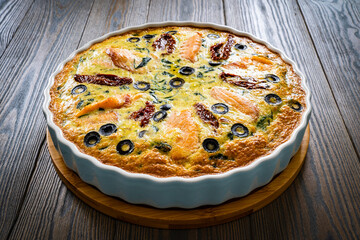 Millet quiche with salmon, spinach and black olives in cooking pan on wooden table
