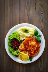 Breaded fried pork chop with potato puree, beetroots, lemon and fresh vegetables on wooden table
