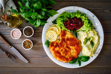 Breaded fried pork chop with potato puree, beetroots, lemon and fresh vegetables on wooden table
