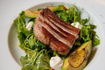 grilled duck steak with fresh vegetables and avocado on a white round plate on a wooden table close up