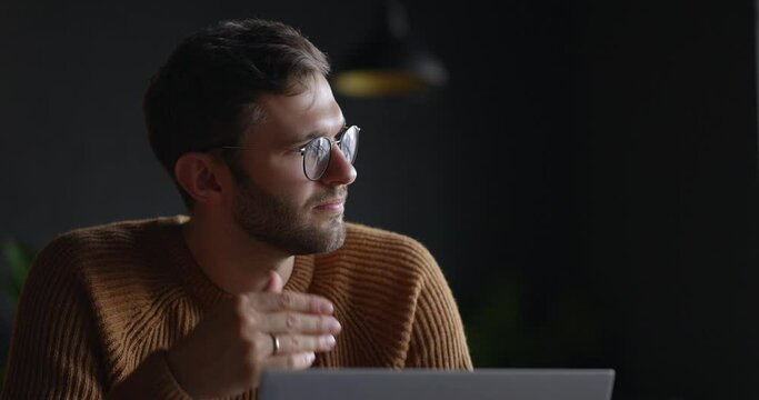 A close-up view of thoughtful serious man in eyeglass sit with laptop thinking of solution at dark office. Slow motion