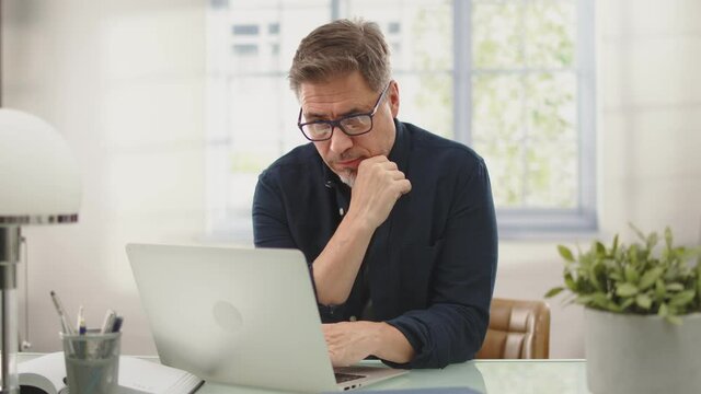 Happy man with laptop computer at home. Businessman working at desk in home office. Portrait of mature age, middle age, mid adult man, bearded, glasses, smiling, thinking, authentic look.