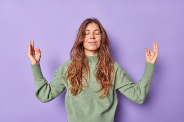 Calm relaxed woman holds hands in zen om gesture keeps eyes closed meditates indoor dressed casually keeps emotions under control peace and patience breathes air freely isolated over purple wall