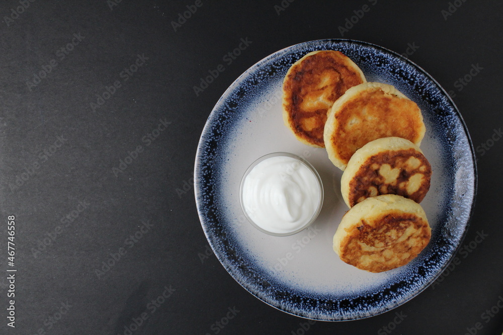 Canvas Prints three cheesecakes lie on a blue plate with a sauce bowl of white sour cream sauce on a black background with space for text. Russian breakfast is traditional. cottage cheese dishes