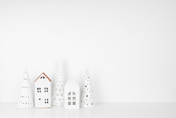 White Christmas decor on a white shelf against a white wall background with copy space. Ceramic trees and village houses.