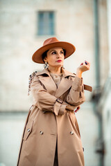 Outdoor fashion portrait of young elegant fashionable brunette woman, model in stylish hat, choker and light raincoat posing at sunset in European city.