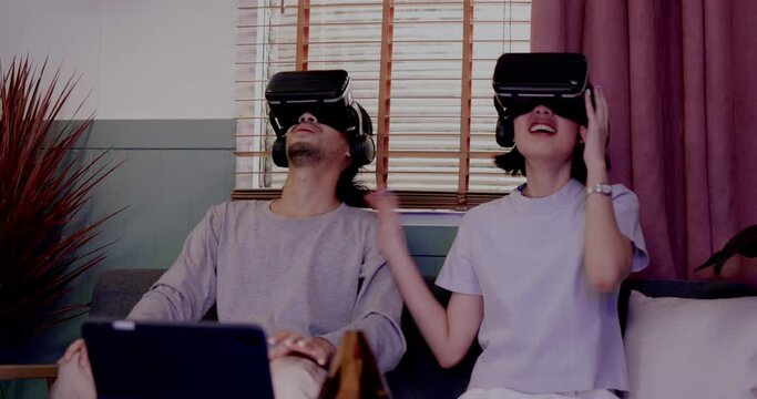 Asian couples wearing modern Virtual reality glasses sit and watch movies in living room at home. Holiday Activity family entertainment technology. living together happiness. Concept metaverse