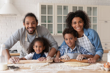 Happy African family baking dessert for dinner together. Sibling children helping couple of parents to cook, rolling dough on table with flour messy, looking at camera, smiling, Head shot portrait