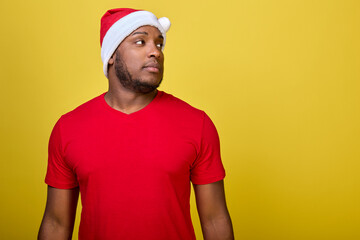 African-American man in a Santa Claus hat and a red T-shirt, standing on a bright yellow background, confidently looks away