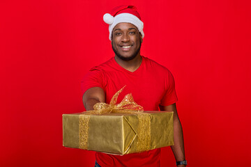 Portrait of a cheerful black guy with a toothy smile in a Santa hat and a red T-shirt giving a Christmas gift in a golden package on a bright red background