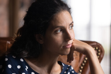 Feeling blue. Pensive serious young hispanic female sit on sofa look aside prop cheek with hand lost in thoughts recall nostalgic moments in memory. Sad thoughtful lady feel melancholy absent minded