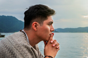 Young man is reflecting while sitting on a dock with water and a mountainous background. Concept of nature