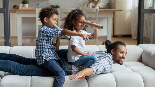 Happy African American Daddy And Little Sibling Kids Playing Funny Active Games On Couch, Children Sailing Dad Like Pirate Boat, Holding Paper Toy Spyglasses. Childhood, Family, Fatherhood