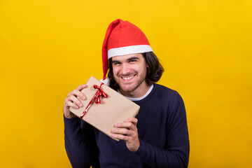 A young man in a Santa Claus hat shows happy Christmas present, a package with brown paper and red ribbon. Yellow background and copy space for your greeting message.