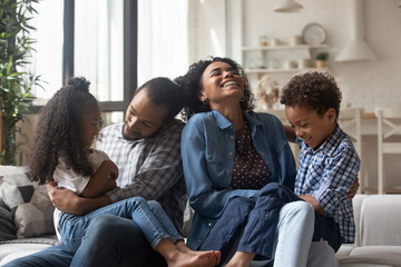 Happy young Black parents cuddling, tickling kids on couch, laughing, having fun, enjoying family...