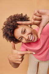 Young positive woman makes camera frame with fingers tilts head and smiles happily searches perfect...