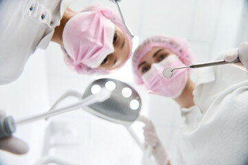 Focus on a stainless steel dental mirror in the hand of a blurred female surgeon dentist adjusting a light beside assistant holding saliva ejector and dental equipment for treating human teeth