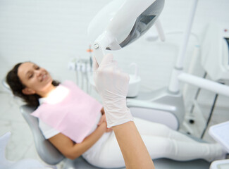 Close-up of a hand in surgical glove adjusting the light against a blurred smiling patient sitting in the dentist's chair and waiting for dental treatment. Medical equipment in dentistry