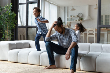 Obraz premium Cheerful hyperactive Black sibling kids running around tired dad, jumping on sofa, playing active games, screaming, laughing. Young father exhausted with watching children, feeling headache, stress