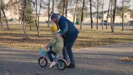 father teaches child ride bike, girl pedals and wheels two-wheeled vehicle, fun horse game city park, happy family, dad and kid relax on day off together, parent entertains the leisure of childhood