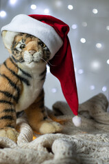 The symbol of 2022 on the Chinese calendar is Tiger. Toy soft plush tiger in a red Santa Claus hat on a glowing background from Christmas garlands.