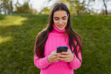 young smiling woman in pink sweater walking in green park using phone