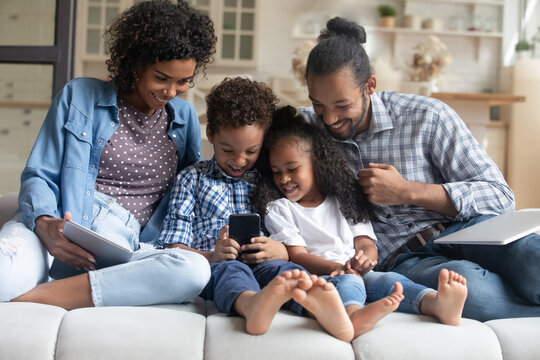Happy African American family couple, little son and daughter using digital gadgets, resting on couch together. Parents watching kid using app on smartphone, playing online game, virtual videogame