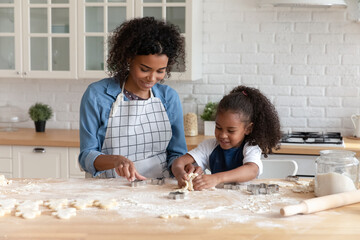Happy African American mom helping cute daughter to bake biscuit, cut dough for cookies. Young...