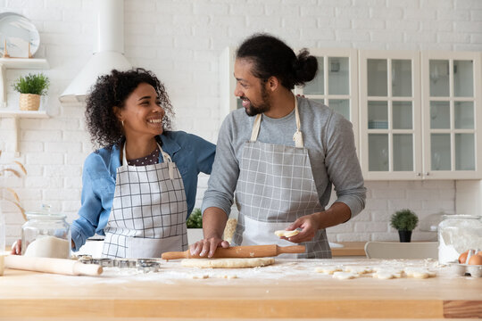 Happy millennial Black couple baking homemade cookies together, rolling dough for biscuit on flour messy tabletop, talking, laughing, having fun, sharing cooking hobby. Food bloggers preparing pastry