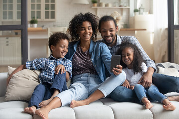 Happy African couple of parents and laughing kids resting on couch holding smartphone, taking selfie on cellphone, making video call, looking at mobile phone screen, smiling, giggling. Family leisure