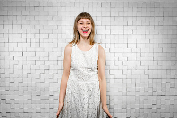 young cheerful caucasian brown-haired woman with bangs in white dress laughs against background of light brick wall with sound insulation. Joyful girl with red lipstick experiences emotions of fun