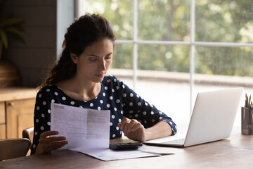 Home banking. Focused young latin female doing paperwork at kitchen think on paying utilities taxes fees check sum on digital calculator. Millennial female read financial documents calculate loan debt