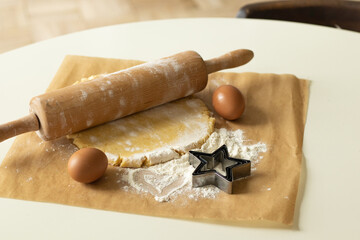 The dough, eggs and flour prepared for cooking lie on parchment paper, next to it lies a wooden rolling pin and an asterisk shape for baking.