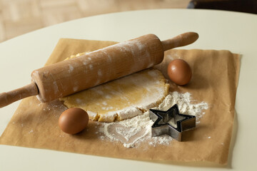 The dough, eggs and flour prepared for cooking lie on parchment paper, next to it lies a wooden rolling pin and an asterisk shape for baking.