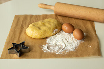 The dough, eggs and flour prepared for cooking lie on parchment paper, next to it is a wooden rolling pin, a heart is drawn on the flour.