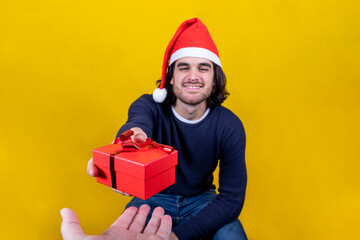 A young man in a Santa Claus hat hands out his Christmas present, a red box with a red ribbon. Yellow background and copy space for your greeting message.