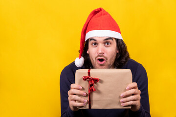 A young man in a Santa Claus hat shows surprised the Christmas present, a package with brown paper and red ribbon. Yellow background and copy space for your greeting message.