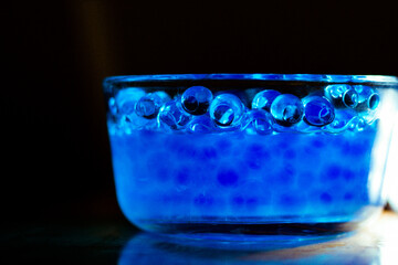 Blue water beads in a clear glass bowl in cold water with a reflection from a light in a  the background.