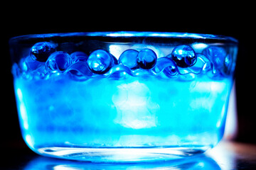 Blue water beads in a clear glass bowl in cold water with a reflection from a light in a  the background.