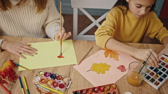 Unknown mother and little daughter drawing autumn leaves with brush and orange watercolor paint, sitting at table. Close up
