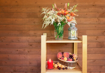 Lilies and astilbe flowers, burning lantern, candle and wicker basket with fruits stand on wooden shelves.