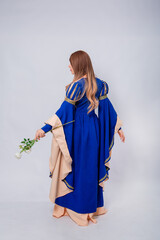 Full-length portrait, back view, attractive woman in medieval, fantasy blue-beige dress with long, flared sleeves posing with a white rose in her hands, isolated on white background.