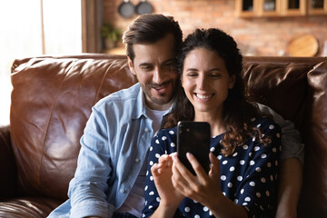 Common interests. Interested young husband cuddle beloved wife on sofa at home watch cute photo video she demonstrate on phone screen. Friendly millennial couple enjoy game at cellphone app read email