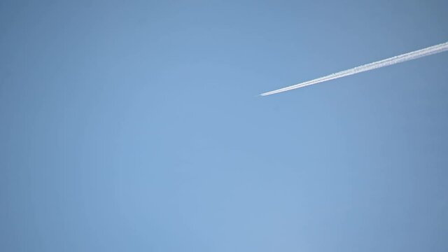High flying airplane jet leaves a contrail. Plane fly against blue cloudless sky leaving water vapor contrails behind as seen from the ground. Aircraft passing by. Real time, static shot
