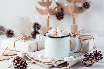 Obraz na płótnie Canvas Cocoa and marshmallows in a mug next to craft gifts against the background of cardboard Christmas trees on a white wooden table. Hot sweet drinks